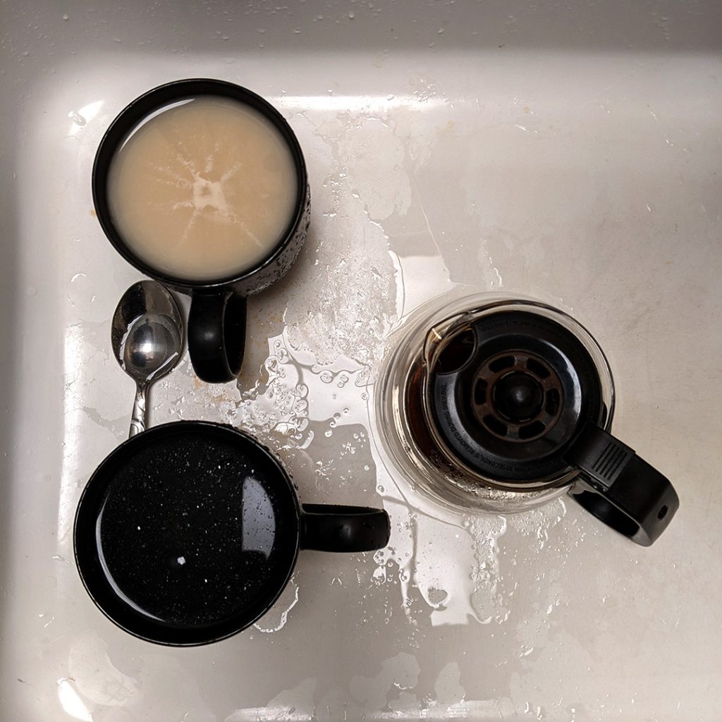 Overhead view of a white sink with dirty coffee items, two black mugs (one with milky residue, one with dark coffee), a metal spoon, and a glass coffee carafe with a black handle, surrounded by water splashes and stains.