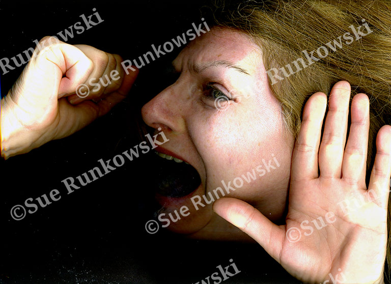 A close-up image of a woman’s face and hands pressed flat against a glass surface, creating a distorted effect. Her expression is one of alarm or distress, with wide eyes and an open mouth as if shouting or gasping. Her long, light brown hair fans out around her face, contributing to the sense of entrapment or urgency. The image conveys a raw, emotional intensity and a sense of being trapped or silenced.