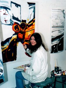 Artist, Sue Runkowski, seated her art school studio, painting a colorful abstract artwork on canvas, surrounded by brushes, paint, and finished prints on the wall.