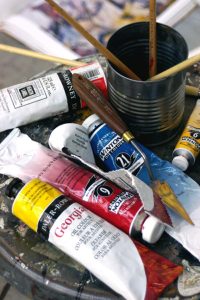Close-up of an artist’s workspace with oil paint tubes, palette knife, paintbrushes, and a metal cup on a paint-splattered surface.