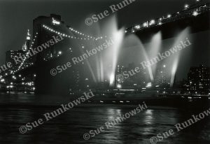 Black-and-white nighttime view of the Brooklyn Bridge illuminated by city lights, with several boats shooting water fountains upward.