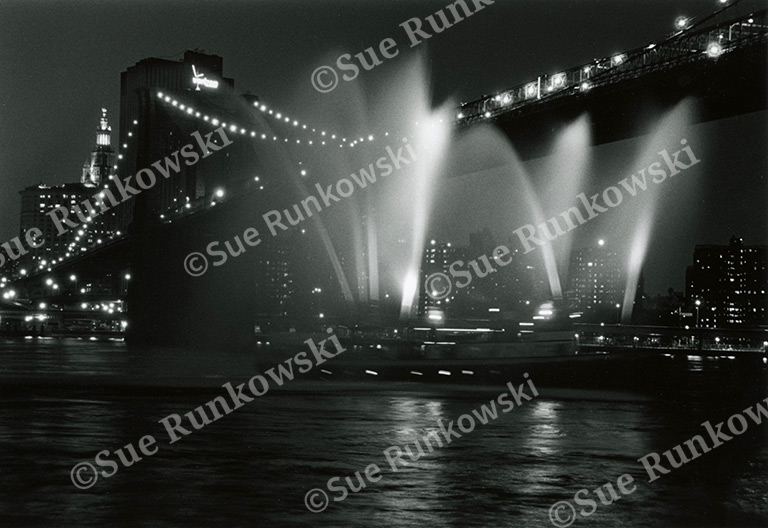 Black-and-white nighttime view of the Brooklyn Bridge illuminated by city lights, with several boats shooting water fountains upward.
