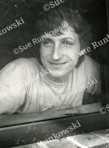 Black-and-white portrait of a young male leaning against a window, captured in soft natural light with a candid expression.