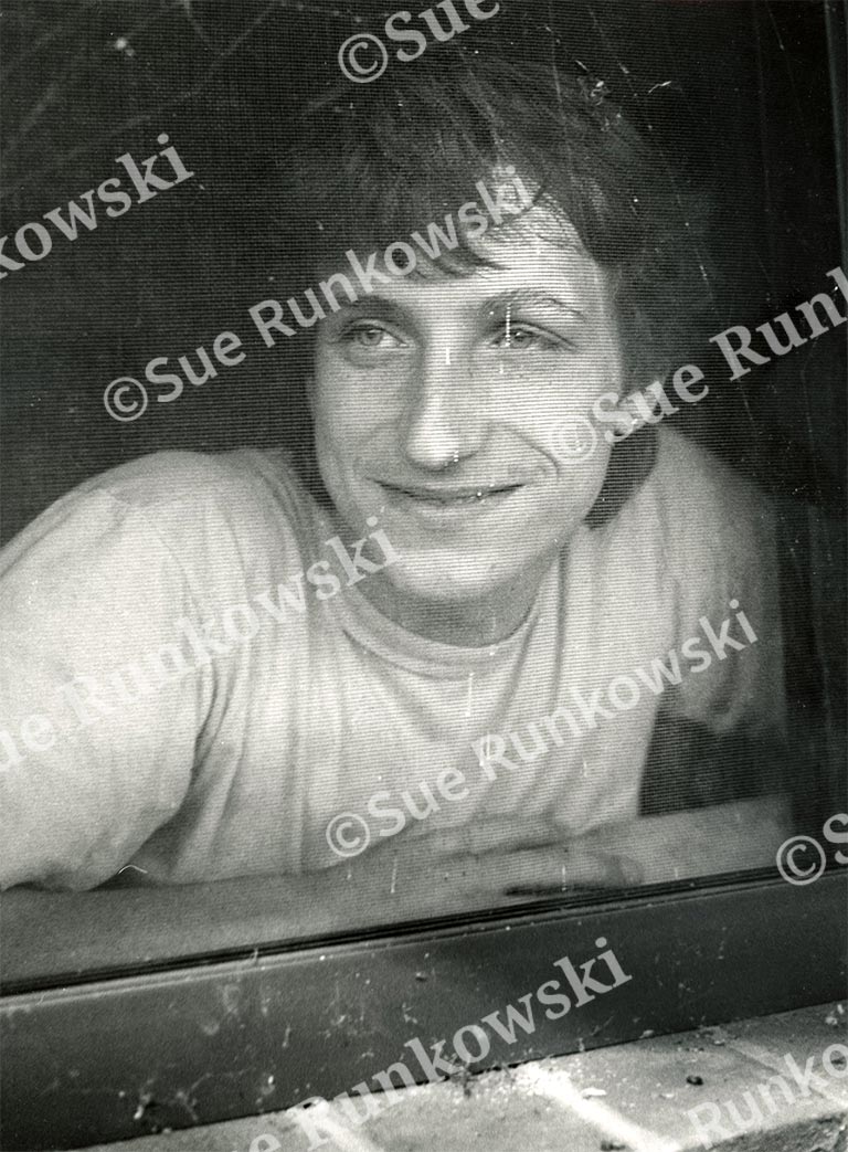 Black-and-white portrait of a young male leaning against a window, captured in soft natural light with a candid expression.