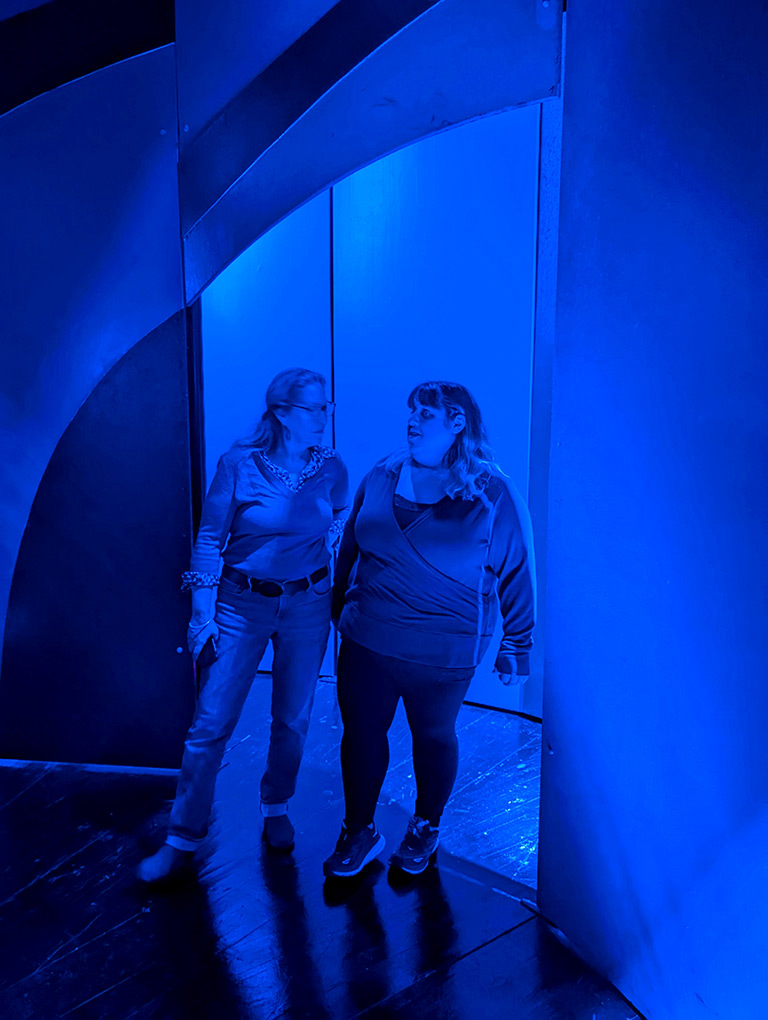 Two women standing in the Hockney Enchanted Tree room showered in blue light