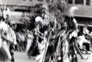 Black and white motion blur photograph of cultural parade dancers, Mexican American Day Parade, traditional costume performance, Aztec-inspired dance regalia, feathered headdresses, dynamic movement, street festival celebration, Indigenous cultural dance, heritage parade procession, urban outdoor event, expressive motion photography, documentary style cultural imagery.