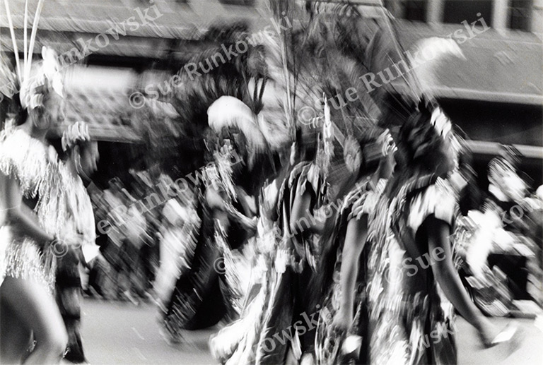 Black and white motion blur photograph of cultural parade dancers, Mexican American Day Parade, traditional costume performance, Aztec-inspired dance regalia, feathered headdresses, dynamic movement, street festival celebration, Indigenous cultural dance, heritage parade procession, urban outdoor event, expressive motion photography, documentary style cultural imagery.