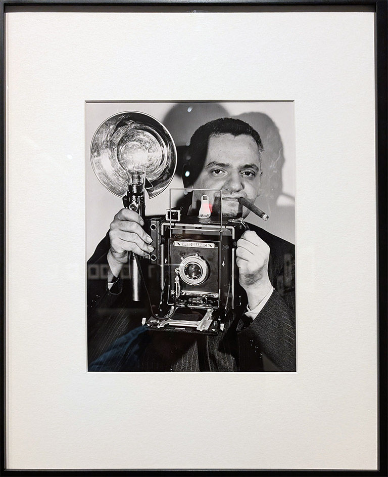 Black-and-white vintage photography self-portrait of photographer Weegee holding a large-format camera and flash, symbolizing classic photojournalism, 1950s street photography, and documentary photography history.