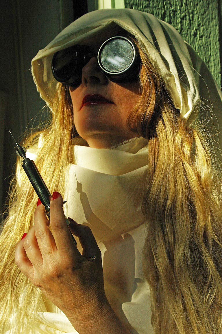 Moody cinematic portrait of a hooded person wearing dark goggles, steampunk goggles, mysterious figure holding a syringe, laboratory syringe prop, dramatic lighting, long blonde hair, white hooded garment, experimental scientist aesthetic, dystopian character portrait, surreal conceptual photography, sci-fi costume, shadowy indoor setting, vintage medical theme, unsettling atmospheric artwork.