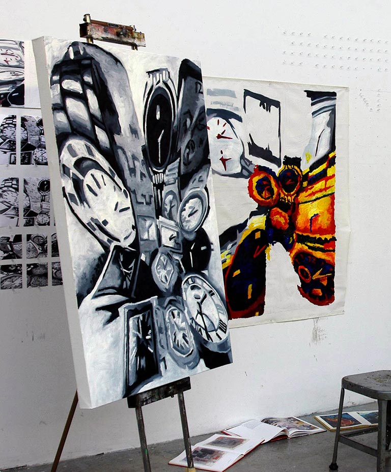 An art studio scene with two large paintings displayed on easels against a white wall. The painting in the foreground is mostly black, white, and gray, filled with overlapping clock faces and abstract mechanical forms. Behind it, a second painting features a bold, symmetrical image resembling a moth or butterfly in orange, yellow, red, and black. Books and printed pages lie open on the floor nearby, and a small stool sits to the right.
