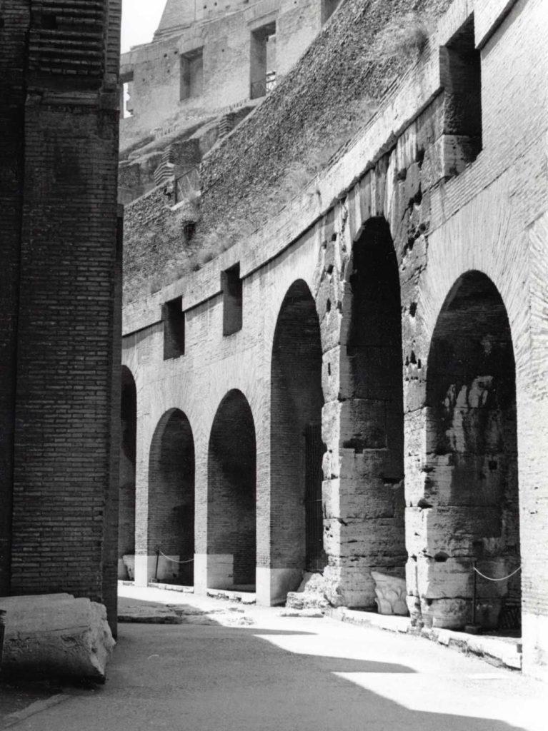 Black-and-white photograph of the interior corridor of the Roman Colosseum. The upper tiers of the amphitheater are partially visible above, evoking a sense of monumental scale and historical grandeur.