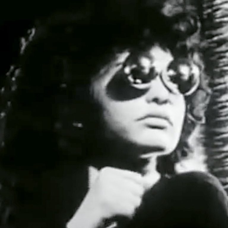 Black-and-white image of a person wearing large round sunglasses and a dark outfit, sitting indoors near a window with light streaming in. The person has curly hair and an introspective expression, with one hand raised near their chin, creating a moody, cinematic atmosphere.