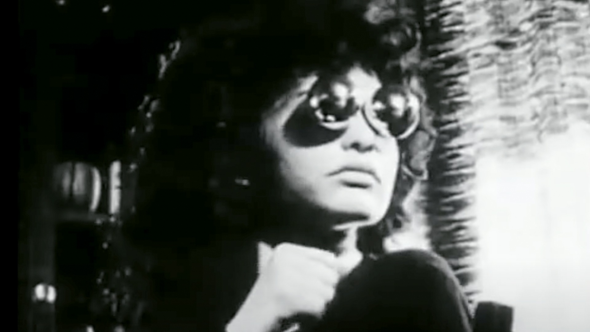 Black-and-white image of a person wearing large round sunglasses and a dark outfit, sitting indoors near a window with light streaming in. The person has curly hair and an introspective expression, with one hand raised near their chin, creating a moody, cinematic atmosphere.