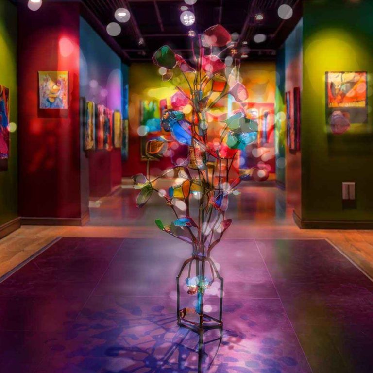 A brightly lit art gallery with colorful walls displaying abstract paintings, centered on a whimsical sculptural installation resembling a glowing tree made of multicolored glass or lights, with soft, floating light reflections throughout the space.