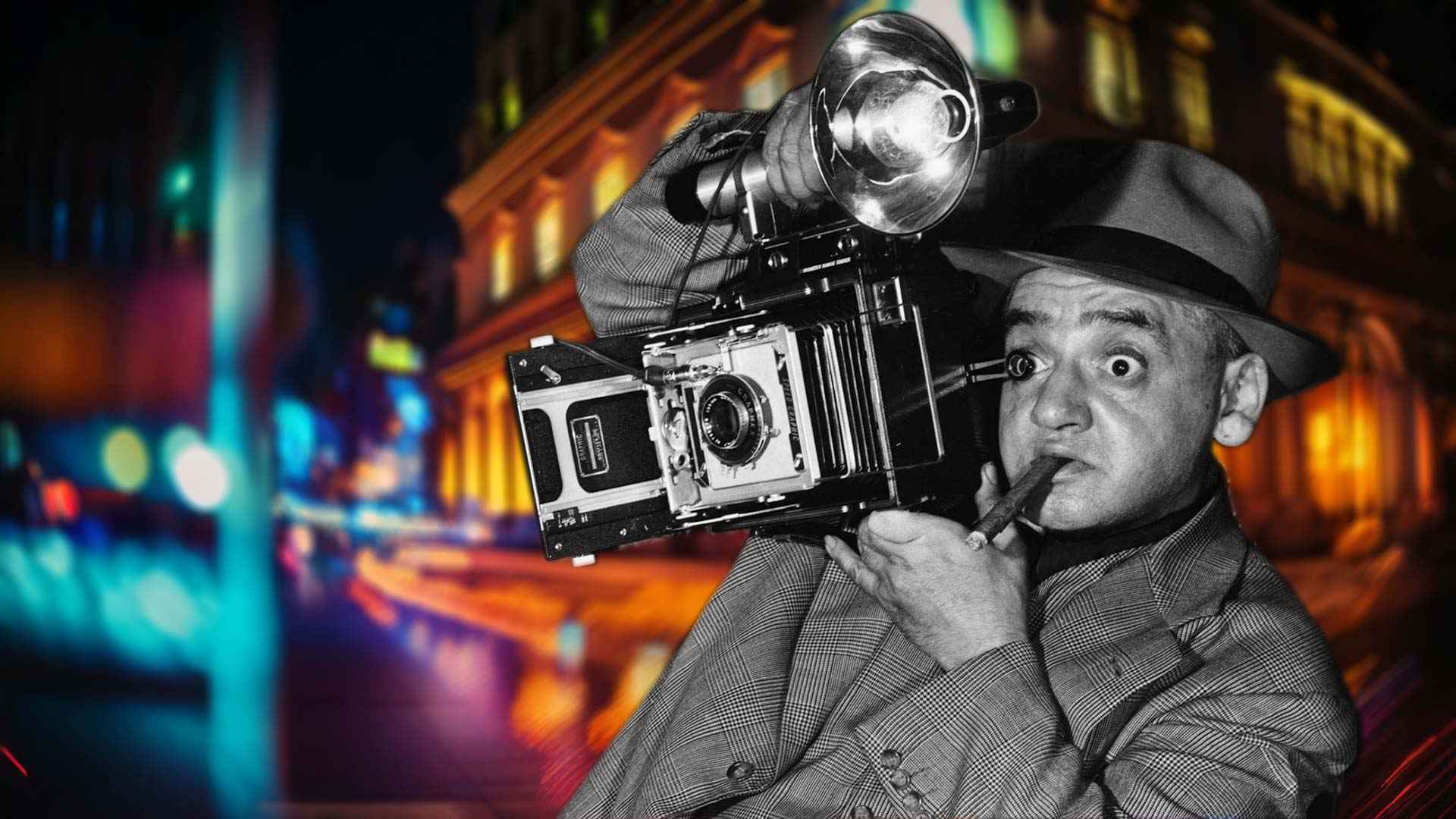 A dramatic, stylized image of Arthur Fellig, aka Weegee, wearing a fedora and suit, holding a large field camera with a flash bulb raised to his eye, a cigar clenched in his mouth. He is shown in black and white against a blurred, colorful nighttime city street with glowing lights and buildings.