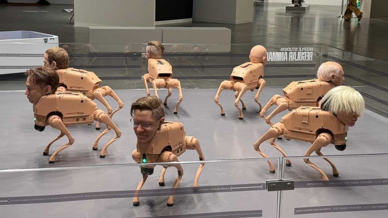 An art installation featuring multiple beige, four-legged robotic dog sculptures arranged in a gallery space, each topped with a realistic human head. The robots stand and move in different directions within a glass-enclosed area on a polished floor, creating a surreal contrast between mechanical bodies and lifelike faces.