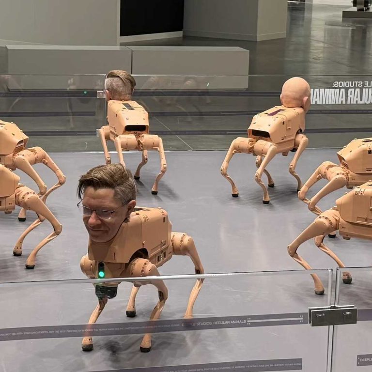 An art installation featuring multiple beige, four-legged robotic dog sculptures arranged in a gallery space, each topped with a realistic human head. The robots stand and move in different directions within a glass-enclosed area on a polished floor, creating a surreal contrast between mechanical bodies and lifelike faces.