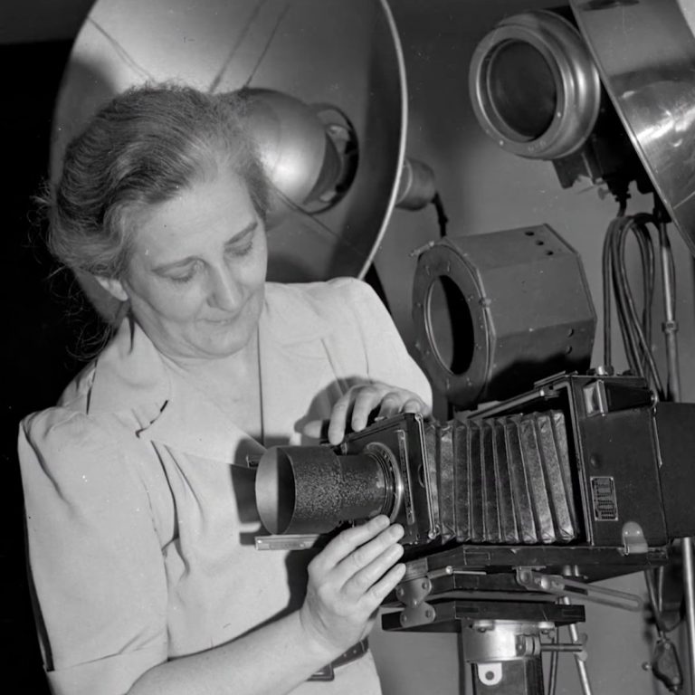 Black-and-white photograph of a woman photographer adjusting a large-format bellows camera on a tripod inside a studio, surrounded by vintage studio lights. She looks down with focused concentration as she fine-tunes the lens, capturing a moment of careful craftsmanship and early photographic practice.