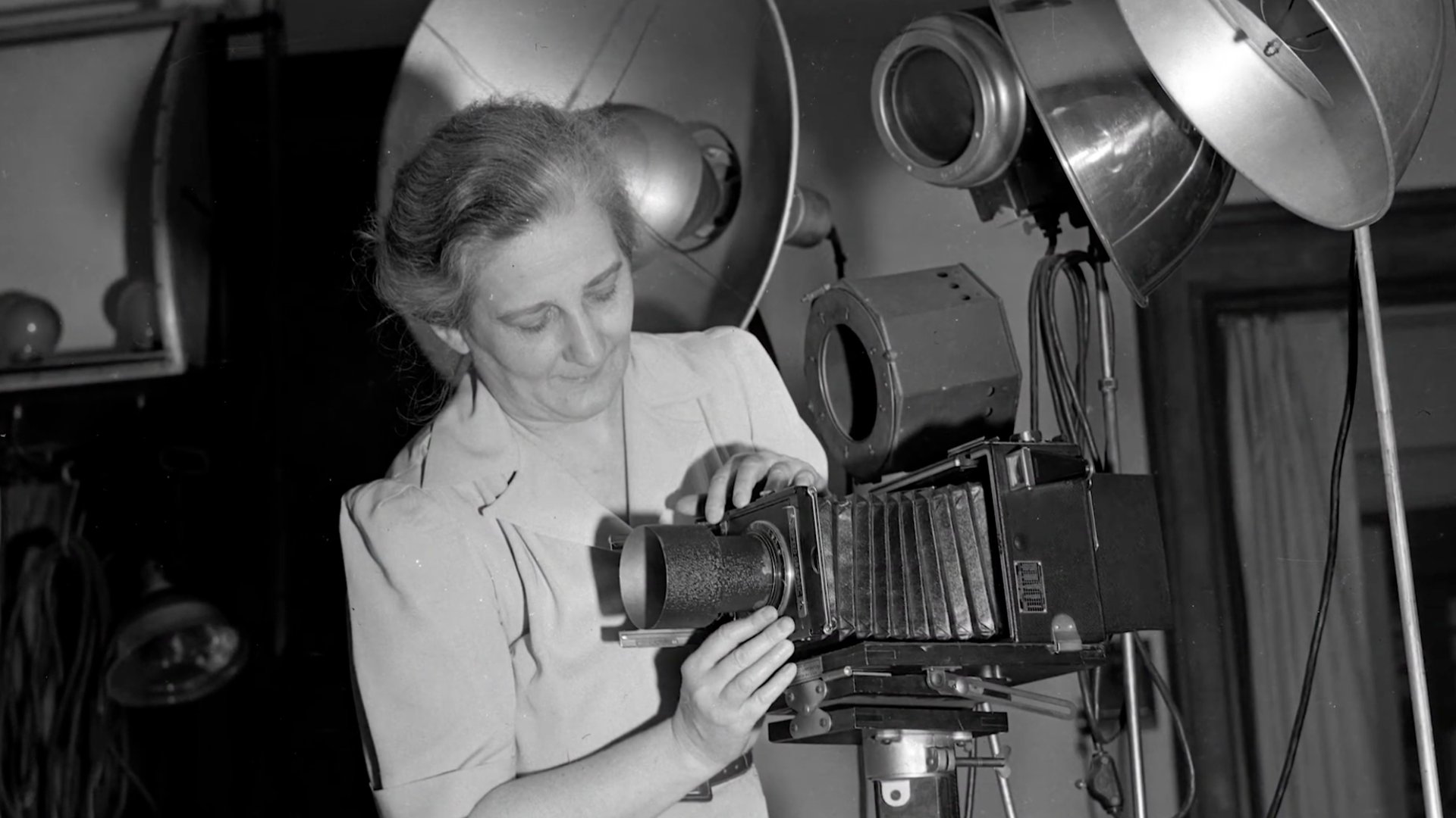 Black-and-white photograph of a woman photographer adjusting a large-format bellows camera on a tripod inside a studio, surrounded by vintage studio lights. She looks down with focused concentration as she fine-tunes the lens, capturing a moment of careful craftsmanship and early photographic practice.