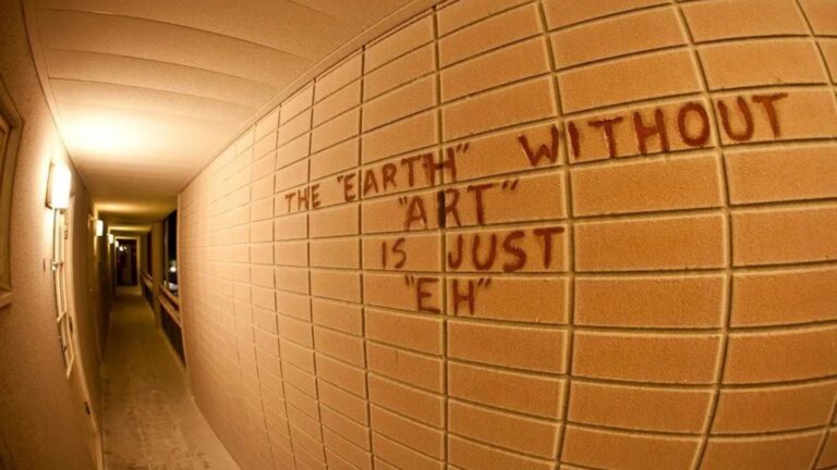 A fisheye view down a narrow hallway with tan brick walls, where red graffiti reads The Earth without Art is just Eh, stretching along the wall under warm overhead lights.