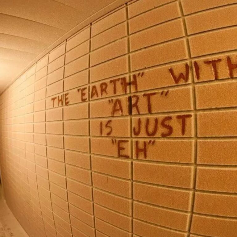 A fisheye view down a narrow hallway with tan brick walls, where red graffiti reads The Earth without Art is just Eh, stretching along the wall under warm overhead lights.