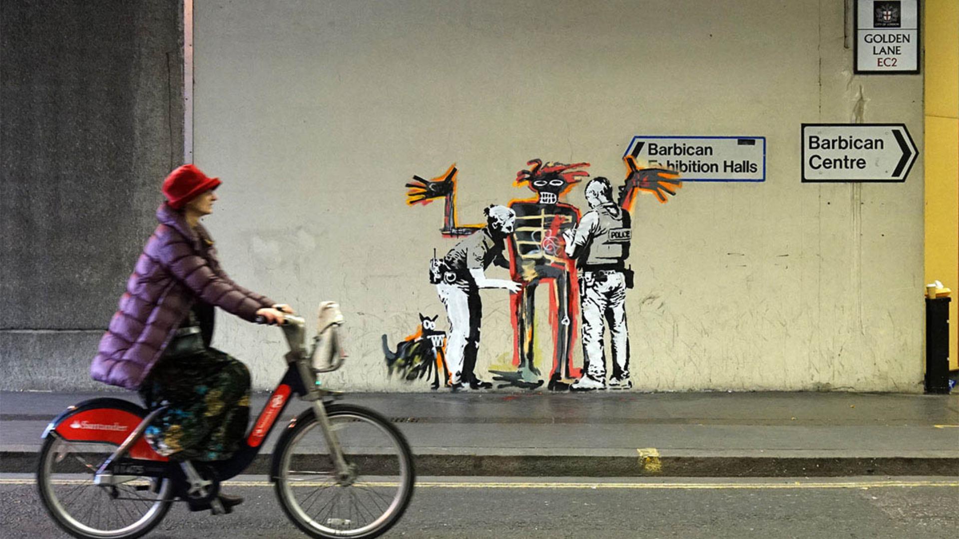 A person in a red hat rides a bicycle past a Banksy mural in London showing two police officers frisking a skeletal, crown-topped figure in the style of Jean-Michel Basquiat, while a small black dog stands nearby and street signs point toward the Barbican Centre.