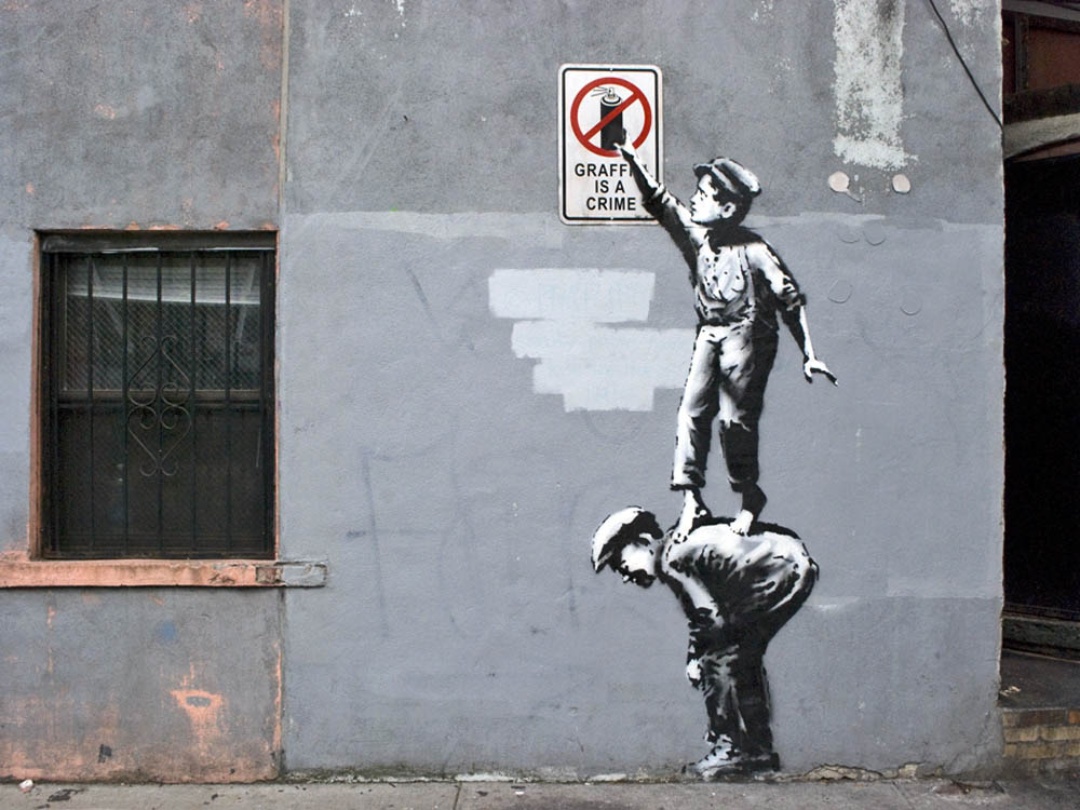 A stencil-style mural on a gray urban wall shows two figures: an adult man bent forward and a child standing on his back to reach a posted sign. The child is holding a spray can up toward a sign that reads “Graffiti is a crime,” with a red prohibition symbol over a spray can. A barred window sits to the left, and the wall shows patches of paint and wear.