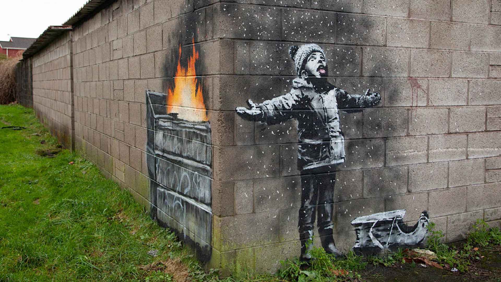 A stencil-style street artwork on a concrete block wall shows a young child wearing a winter hat and coat, standing with arms outstretched and tongue out as if catching falling snow. White specks surround the child like snowflakes. Around the corner of the wall, a painted dumpster is on fire, revealing that the “snow” is actually ash drifting from the flames. Grass and a narrow path run alongside the wall.