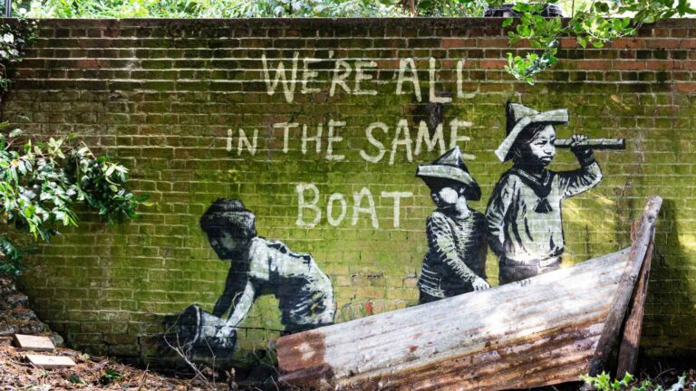 A weathered brick wall covered in green moss features a stencil-style mural of three children in a makeshift boat made from a rusted metal sheet. One child looks through a telescope, another sits behind, and a third appears to bail water with a bucket. Above them, painted in white, are the words: “We’re all in the same boat.” Overgrown plants and dappled sunlight frame the scene.