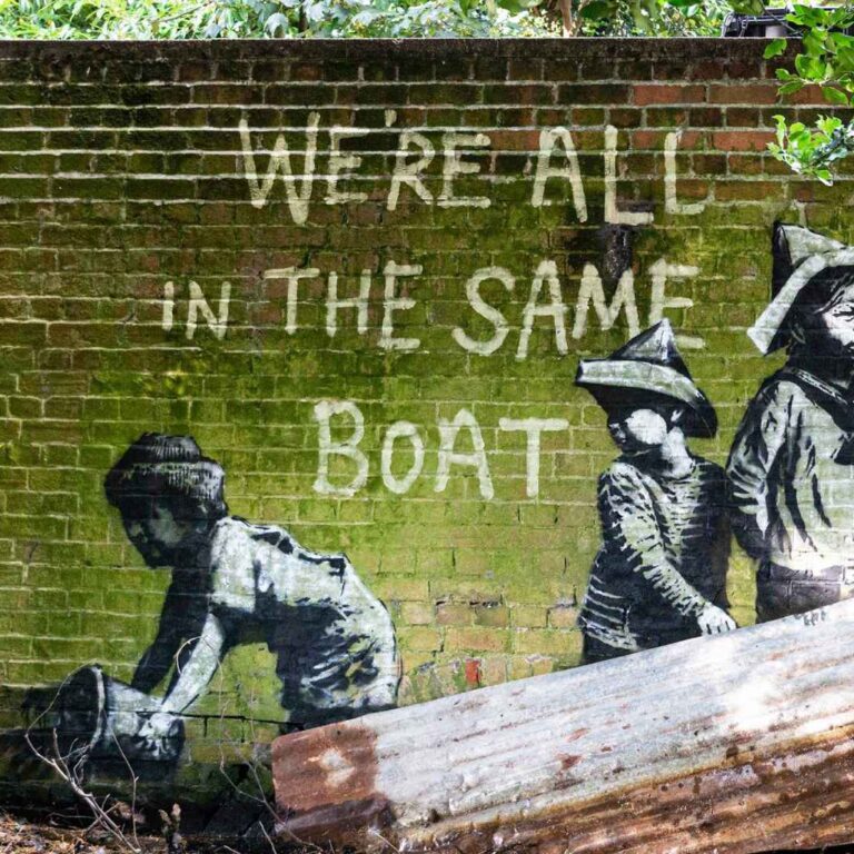 A weathered brick wall covered in green moss features a stencil-style mural of three children in a makeshift boat made from a rusted metal sheet. One child looks through a telescope, another sits behind, and a third appears to bail water with a bucket. Above them, painted in white, are the words: “We’re all in the same boat.” Overgrown plants and dappled sunlight frame the scene.