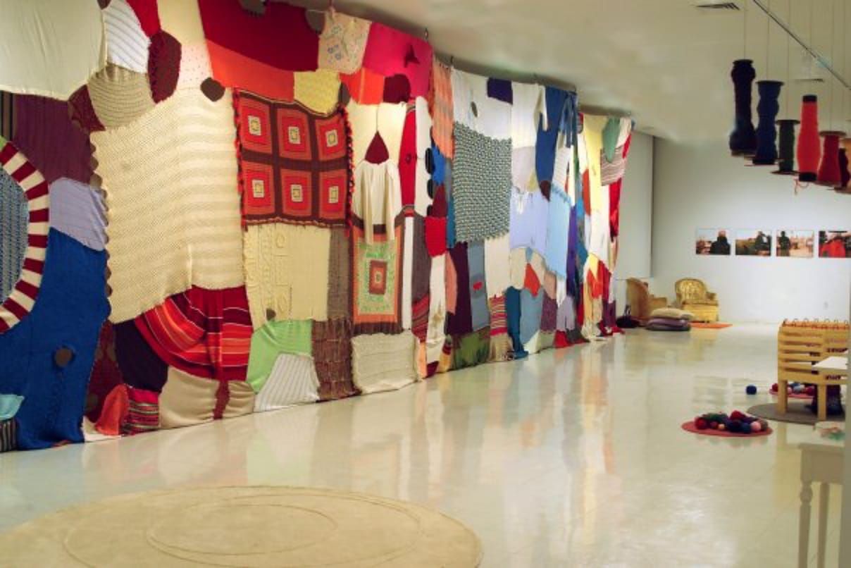 A large gallery installation made from stitched-together sweaters, blankets, and knitted panels forms a colorful patchwork wall, with yarn, stools, and handmade objects arranged on the floor and small photographs displayed on the far wall.