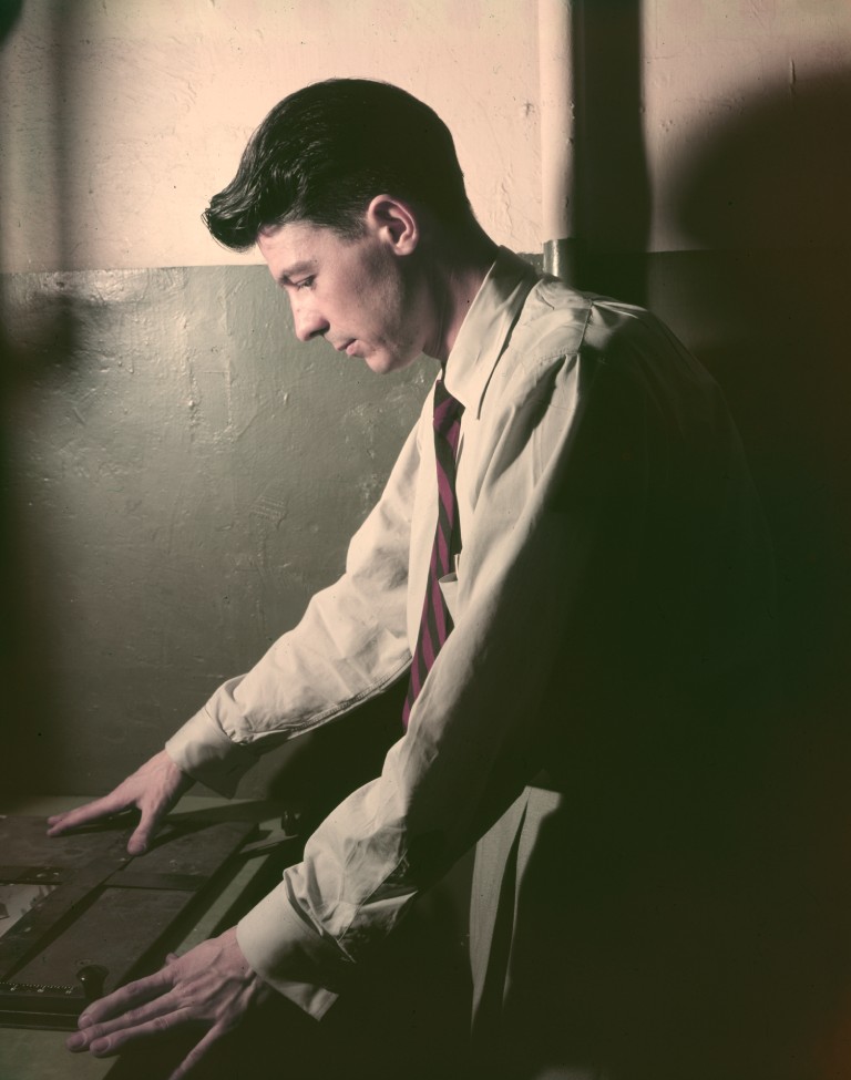 A softly lit portrait shows a young man standing in profile in a dim darkroom, leaning over a work surface with both hands resting near photographic trays or prints. He wears a light-colored button-down shirt and a striped tie, with his dark hair neatly combed back. His expression is focused and contemplative as he looks downward at the developing photographs. The moody lighting and shadows create an intimate, nostalgic atmosphere, emphasizing the quiet concentration of the photographic process.