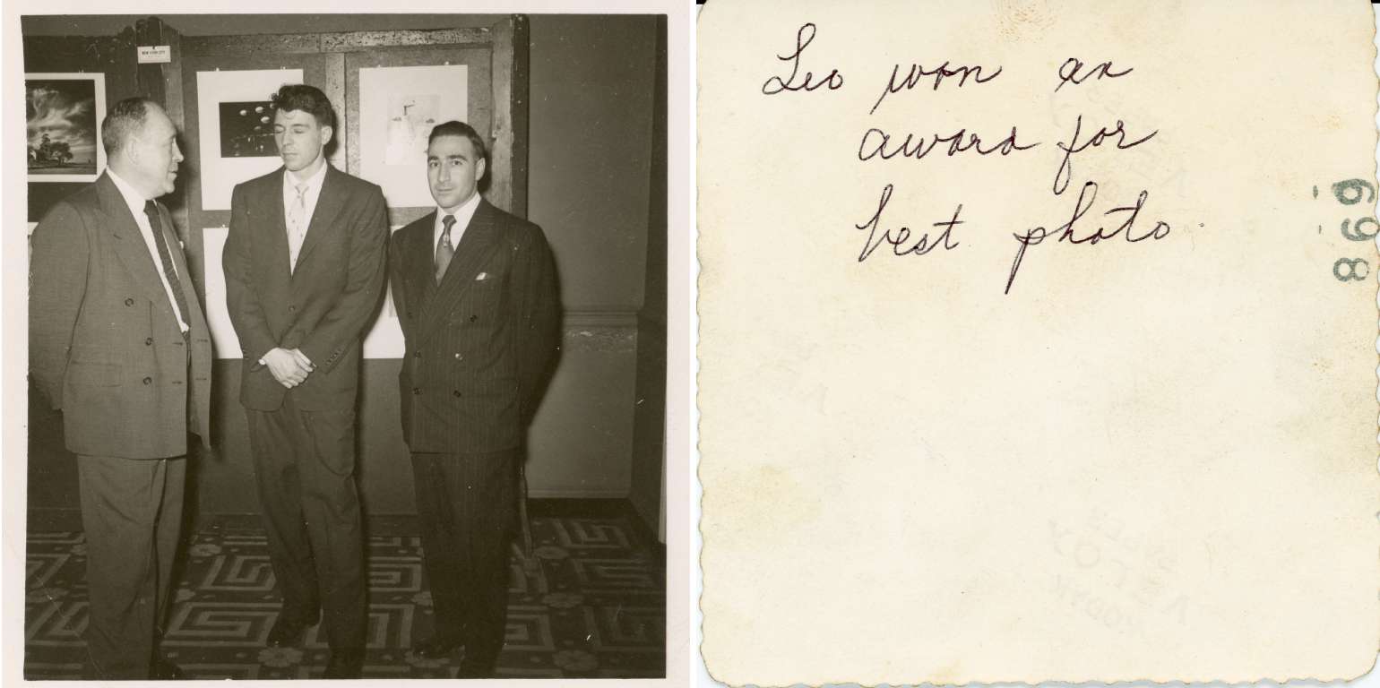 Black-and-white photograph of three men wearing suits standing indoors in front of a wall displaying framed photographs. One man looks toward another while the third faces the camera. The setting appears to be an art or photography exhibition, with patterned flooring and mounted prints visible in the background.