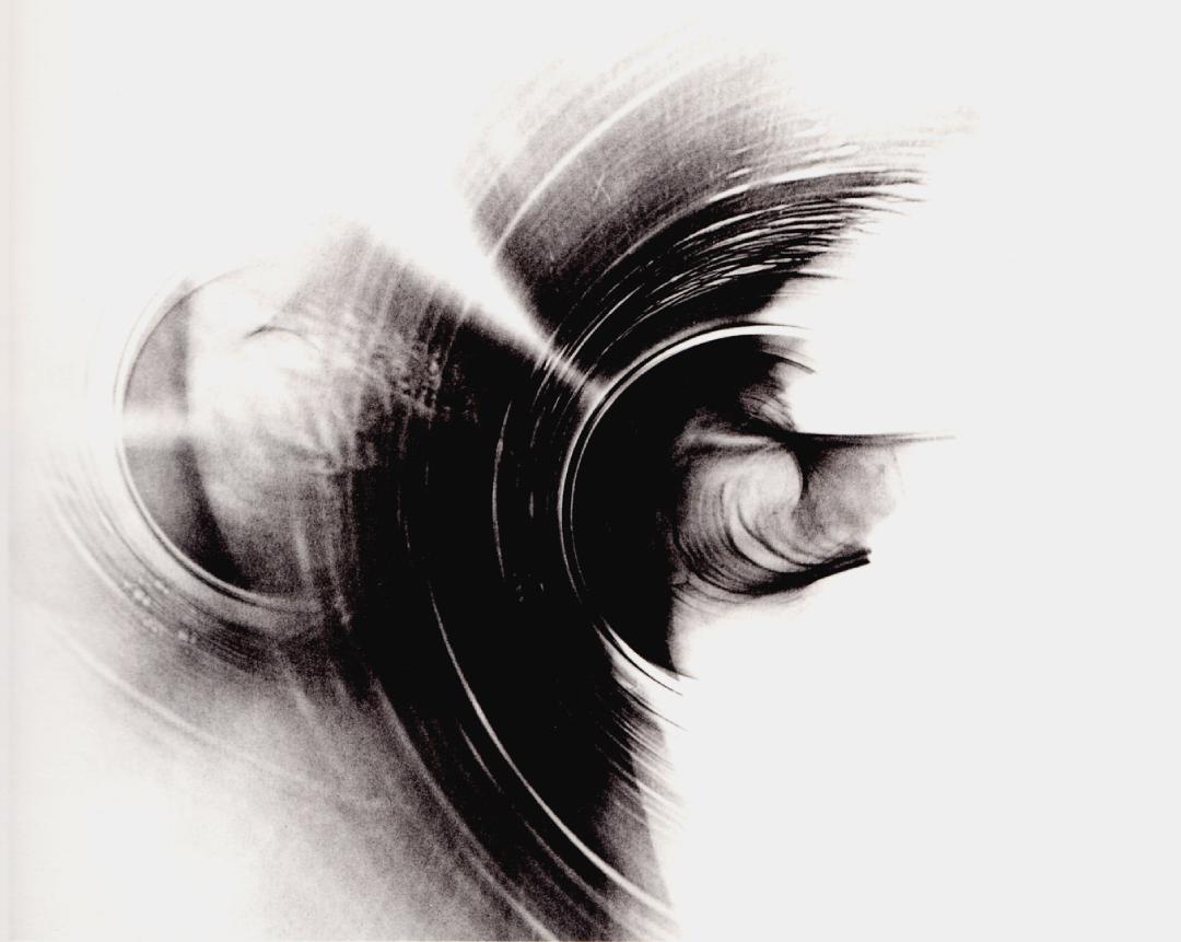 An abstract black-and-white photogenic image composed of sweeping circular forms, blurred arcs, and luminous streaks of light against a bright background. The composition resembles spinning machinery, celestial motion, or flowing energy waves, creating a dynamic sense of movement and surreal optical depth.